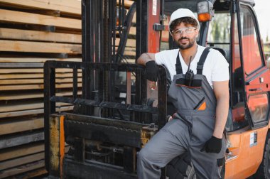 Forklifte yaslanmış, arka planda kereste yığınları olan gülümseyen bir depo işçisinin portresi kereste endüstrisini gözler önüne seriyor.