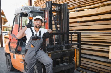Depo işçisi, bir kereste deposundaki kereste yığınının yanında bir forklifte yaslanırken baş parmak işareti yapıyor.