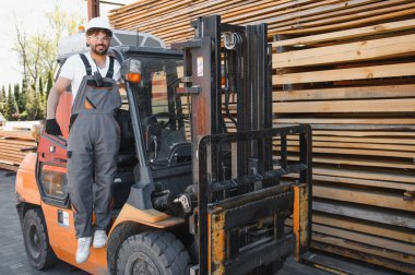 Güvenlik kaskı takan depo çalışanı ahşap bir depodaki kereste yığınlarının yanında bir forkliftin üzerinde iş tulumu giyiyor.