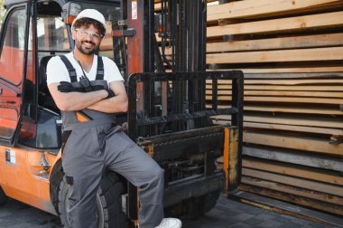 Kereste deposunda forklifte yaslanmış, kollarını kavuşturmuş, iş kıyafetleri içinde gülümseyen bir işçinin portresi.