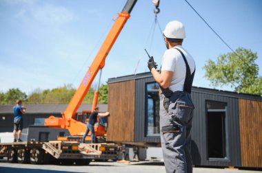 İnşaat işçisi telsiz kullanıyor, vinç kaldırma modüler evini kamyona yüklüyor, verimli prefabrik konut taşımacılığı sergiliyor.