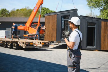 Mühendis, prefabrik modüler bir evin bir kamyona yüklenmesini denetlemek için uzaktan kumanda kullanıyor.