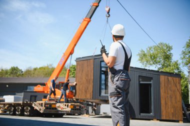 İnşaat işçisi telsiz kullanıyor, vinç kaldırma modüler evini denetliyor, yenilikçi konut çözümleri var.