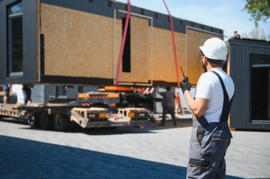 İnşaat işçisi telsiz kullanıyor. İnşaat alanındaki kamyona prefabrik ev bölümünün yüklenmesini koordine ediyor.