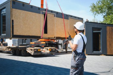 Mühendis prefabrik modüler bir evin taşınmasını denetliyor, vinç operasyonunu telsizle koordine ediyor.