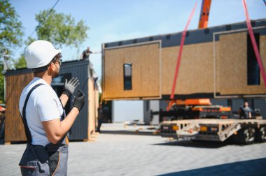 İnşaat işçisi telsiz kullanıyor, prefabrik ev bölümünü kamyona yüklüyor, modern konut inşaatında verimlilik gösteriyor.