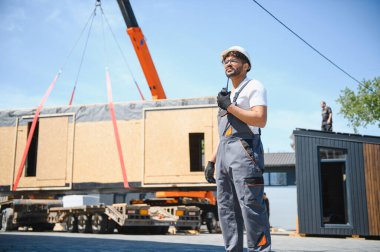 İnşaat işçisi vinç kaldırırken telsiz kullanıyor prefabrik ev modülünü kamyona yüklüyor, yenilikçi bina teknolojisi.