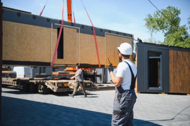 Mühendis telsiz kullanıyor, vinç koordine ederek prefabrik ev bölümünü modüler inşaat alanındaki bir kamyona yüklüyor.