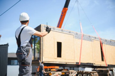 İnşaat işçisi telsiz kullanıyor, vinç kaldırma, prefabrik modüler evi inşaat alanındaki kamyona yönlendiriyor.