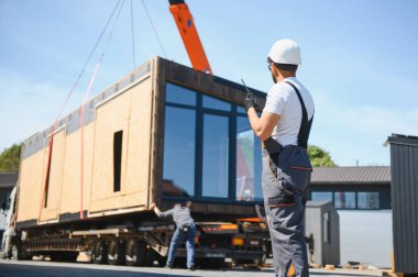 İnşaat işçisi, prefabrik ev modülünü kamyona yüklemeyi koordine eden telsiz kullanıyor. Hızlı ve verimli bir inşaat teknolojisi sağlıyor.