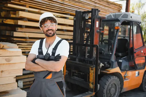 Kendine güvenen Arap ahşap işçisi kollarını kavuşturup gülümsüyor, güvenlik kaskı ve telsizi takıyor, forklift ve ambardaki kereste yığınlarının önünde duruyor.