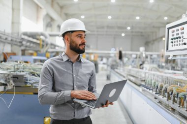 Endüstriyel mühendis bilgisayar kullanıyor, işletmeleri yönetiyor ve büyük modern fabrikada makineleri denetliyor.