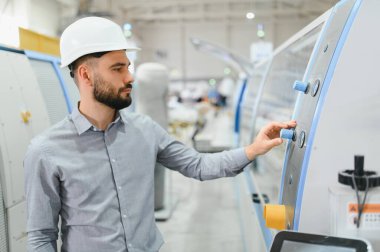 Modern fabrikada sabit kasklı çalışma makineleri takan endüstri mühendisi kontrolleri ayarlıyor ve üretim sürecini izliyor