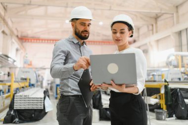 Şapkalı iki mühendis bir dizüstü bilgisayar ve pano kullanıyor. Fabrika ortamında iş konuşuyorlar.