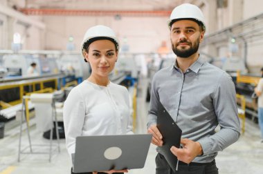 Dizüstü bilgisayar ve pano tutan iki mühendis modern bir fabrikanın üretim hattını kontrol ediyorlar.