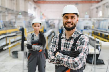 Modern bir fabrikada çapraz silahlarla poz veren gülümseyen iki sanayi işçisinin portresi, ekip çalışması ve imalat alanında uzmanlık gösteriyor.