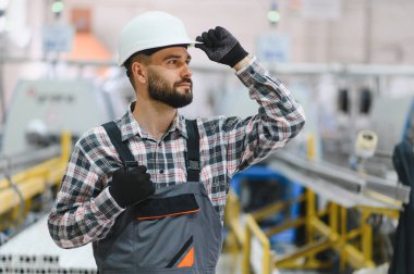 Sanayi mühendisi, yoğun bir imalat fabrikasında çalışırken kaskını düzeltiyor, güvenlik ve üretim süreçlerini denetliyor.