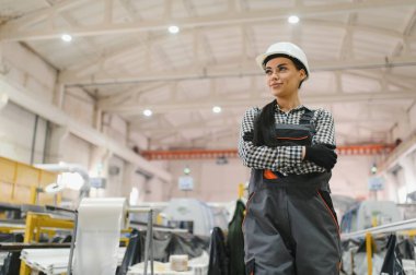 İş kıyafetleri ve şapka giyen kendine güvenen kadın yönetici, büyük modern bir fabrikada çapraz kollarla üretim yapıyor.