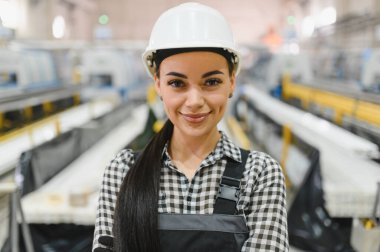 Kendine güvenen kadın fabrika işçisi güvenlik kaskı takıyor ve endüstriyel üretim hattında makinelerin önünde gülümsüyor.