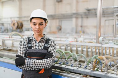 Fabrikada üretim bandının önünde kollarını çapraz bağlamış duran kasklı ve üniformalı kadın endüstriyel işçiler.