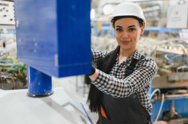 Kendine güvenen kadın fabrika işçisi koruyucu giysiler giyerken ve iş güvenliğini sağlarken ağır endüstriyel makineler kullanıyor.