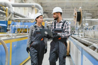 Tulumlu ve şapkalı iki fabrika işçisi, modern bir endüstriyel tesiste ellerinde dizüstü bilgisayarlarıyla yürüyüp konuşuyorlar.