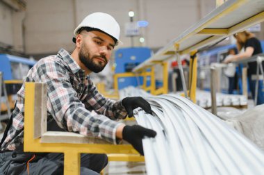 Koruyucu eldiven ve miğfer takan fabrika işçisi pencere ve kapı üretim tesisinde alüminyum profilleri dikkatle inceliyor.