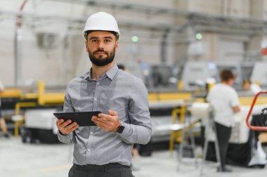 Endüstriyel mühendis kasket takıyor ve tablet tutuyor, operasyonları yönetiyor ve modern fabrikada üretim sürecini denetliyor.