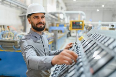 Modern fabrikada sabit kasklı çalışan makineler takan endüstri mühendisi kontrol panelindeki düğmelere basıyor.