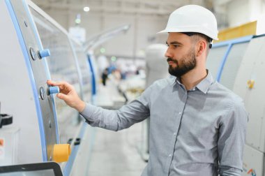 Endüstriyel mühendis sabit bir şapka takıyor, makine kontrol panelini ayarlarken aynı zamanda modern bir üretim tesisinde operasyonları denetliyor.