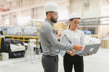 Kasketli iki mühendis büyük bir fabrikada iş konuşmak için dizüstü bilgisayar ve dosya kullanıyor.