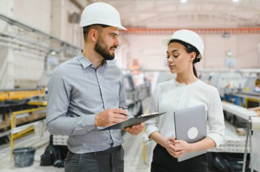 İki mühendis, bir erkek ve bir kadın, bir fabrikada üretimi tartışıyor, bir imalat ortamında takım çalışması, uzmanlık ve profesyonellik gösteriyorlar.