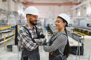 Sert şapkalar ve tulumlar giyen iki fabrika işçisi çapraz kollarla büyük bir endüstriyel üretim alanında birbirlerine gülümsüyorlar.