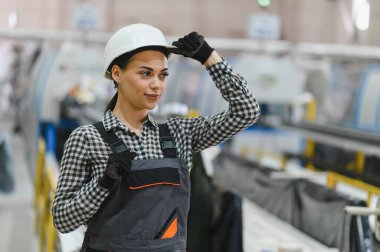Güçlü bir fabrika ortamında gelişmiş makinelerle çevriliyken kaskını düzelten kendine güvenen kadın sanayi işçisi.