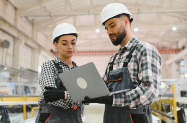 Güvenlik kaskı takan ve dizüstü bilgisayar kullanan iki fabrika işçisi modern bir fabrikada endüstriyel işlemleri yönetiyor.