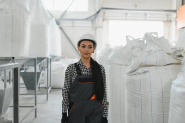 Kendine güvenen kadın mühendis beyaz kasketli ve tulumlu gıda işleme tesisinde yürüyor, büyük un çuvallarını inceliyor.