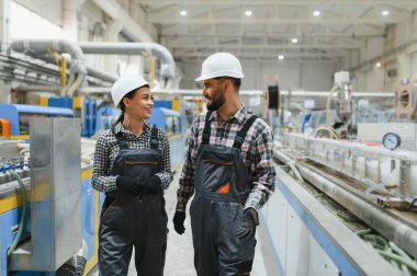 İki fabrika işçisi tulum giyiyor ve kasketli bir şekilde yürüyorlar ve bir üretim bandının yanında konuşuyorlar, gülümsüyorlar ve iş tartışıyorlar.