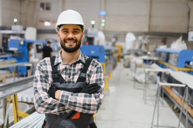 PVC pencere ve kapı üretim fabrikasında kolları çaprazlanmış, kasklı ve tulumlu gülümseyen fabrika işçisi.