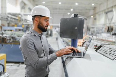 Kask takan endüstri mühendisi, modern fabrikada üretim hattındaki makineleri izlemek ve kontrol etmek, verimliliği optimize etmek ve kaliteyi sağlamak için dizüstü bilgisayar kullanıyor.