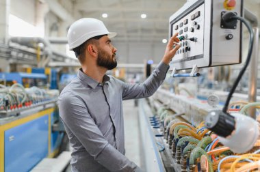 Modern fabrikada sabit kasklı çalışan makineler takan endüstri mühendisi kontrol panelindeki düğmelere basıyor.