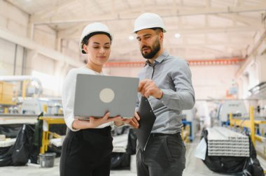 Şapkalı iki mühendis, modern bir fabrika ortamında üretimi tartışmak için dizüstü bilgisayar ve pano kullanıyor.