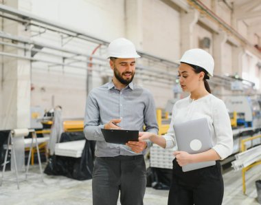 Kasketli iki mühendis modern bir fabrika ortamında bir dosya ve dizüstü bilgisayarda planları gözden geçiriyorlar.
