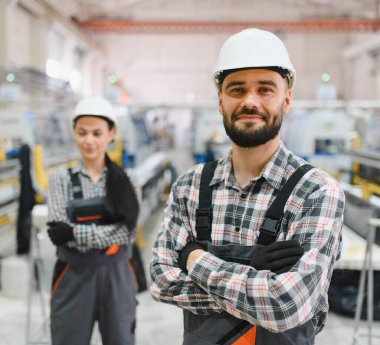 Tulumlu ve kasklı iki fabrika işçisi büyük bir endüstriyel üretim alanında kollarını kavuşturarak gururla ayakta duruyorlar.