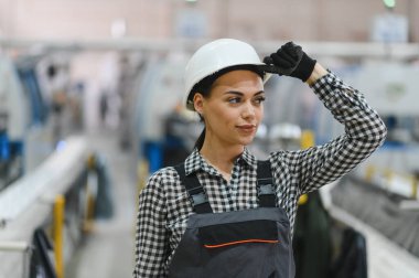 Kendine güvenen kadın fabrika işçisi hareketli bir endüstriyel ortamda şapkasını düzeltiyor, profesyonellik ve güvenlik bilincini gösteriyor.