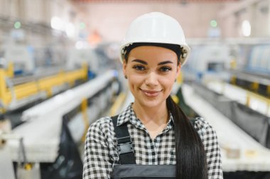 Plastik bir pencere ve kapı imalat fabrikasında kasketli ve tulumlu genç bayan fabrika işçisi güvenle gülümsüyor.
