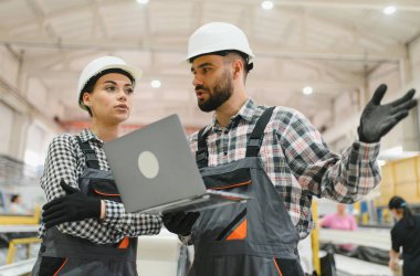 Kask ve tulum giyen iki fabrika işçisi dizüstü bilgisayar kullanıyor ve büyük bir endüstriyel tesiste iş konuşuyorlar.