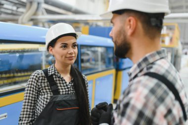 Tulum ve şapka giyen iki fabrika işçisi üretim hattındaki makinelerin yanındaki operasyonları tartışıyorlar.