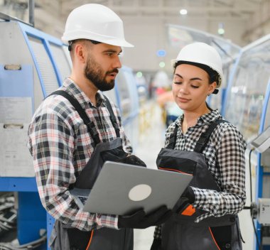 Önlüklü ve şapkalı iki fabrika işçisi bir dizüstü bilgisayar kullanıyor, endüstriyel üretim bandında iş konuşuyorlar.
