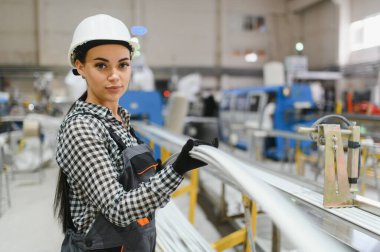 Koruyucu iş kıyafetleri giyen kadın fabrika işçisi ve modern bir fabrikanın üretim hattındaki PVC borularını kontrol ediyor.