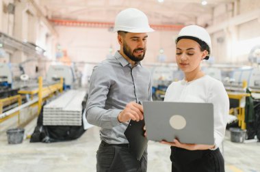 Şapkalı iki mühendis dizüstü bilgisayar kullanıyor ve modern bir fabrika ortamında bir projeyi tartışıyorlar.